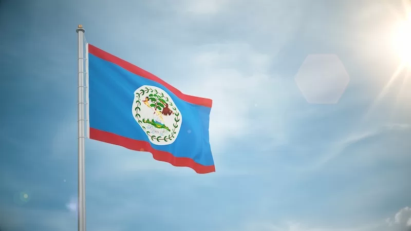 Belize national flag waving on a flagpole against a bright blue sky with sunlight and lens flare