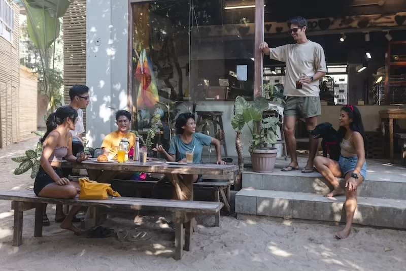 Group of young adults socializing outside a café near the beach, seated around a wooden table with drinks in a relaxed outdoor setting