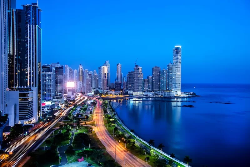 Panama City skyline at night with illuminated towers, coastal boulevard, and light trails running along the waterfront