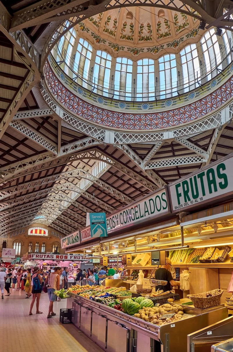 Interior of Valencia’s Central Market with fruit stalls, shoppers, ornate ironwork, and a decorative domed ceiling