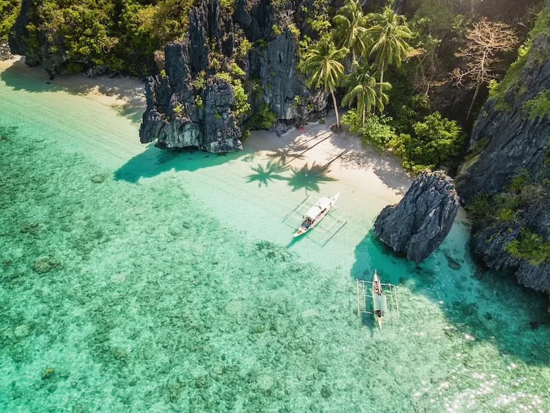 Small outrigger boat floating in a clear turquoise cove surrounded by steep limestone cliffs and a narrow sandy beach