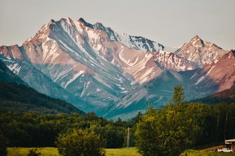 Snow-capped mountain range rising above a forested valley under soft evening light.
