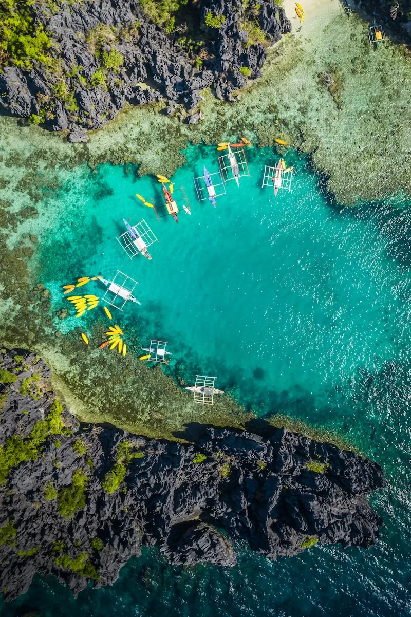 Aerial view of kayaks and small outrigger boats floating in a bright turquoise lagoon bordered by dark rocky cliffs
