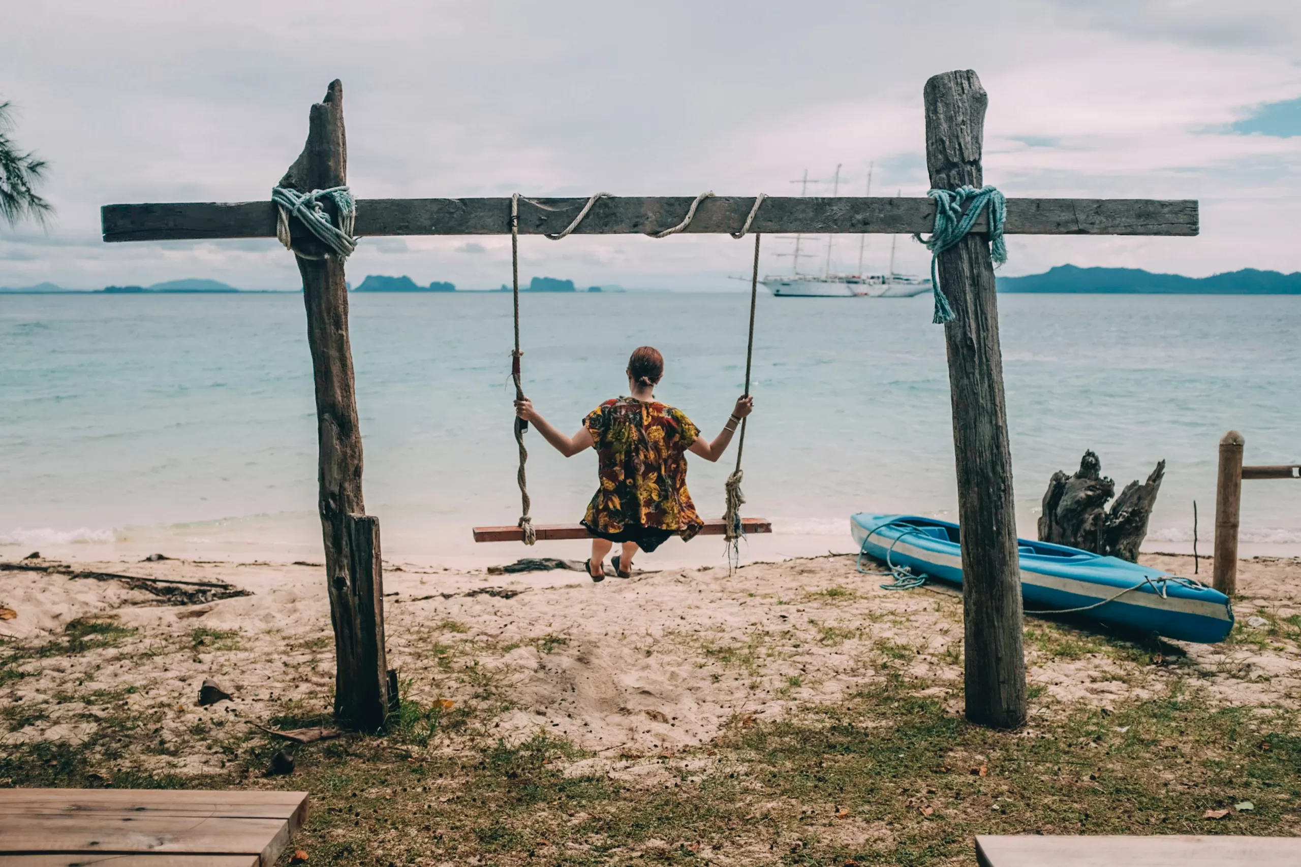 Woman sitting on a rustic wooden swing facing the sea on a quiet tropical beach, with a blue kayak and anchored sailboats in the background