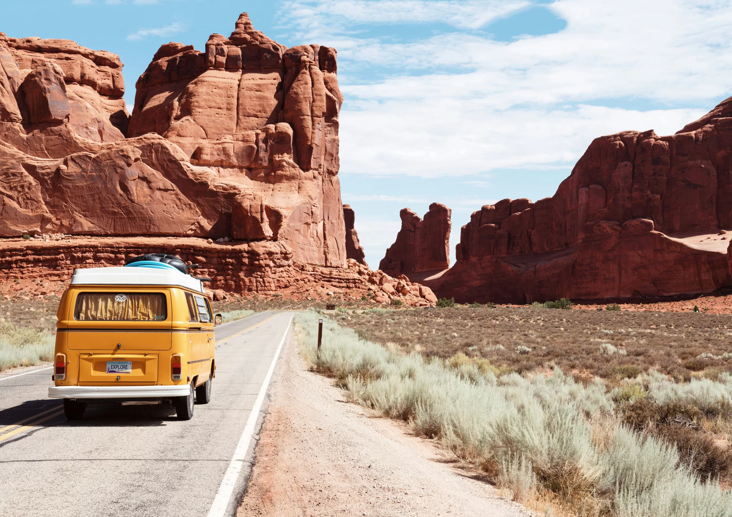 Yellow camper van driving along an open road through a dramatic red rock desert landscape beneath a bright blue sky