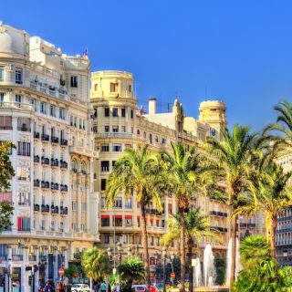 Sunlit view of Valencia, Spain, featuring ornate historic buildings along a palm-lined street, with pedestrians and light traffic under a clear blue sky.