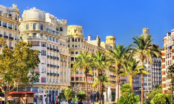 Sunlit view of Valencia, Spain, featuring ornate historic buildings along a palm-lined street, with pedestrians and light traffic under a clear blue sky.
