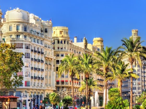 Sunlit view of Valencia, Spain, featuring ornate historic buildings along a palm-lined street, with pedestrians and light traffic under a clear blue sky.