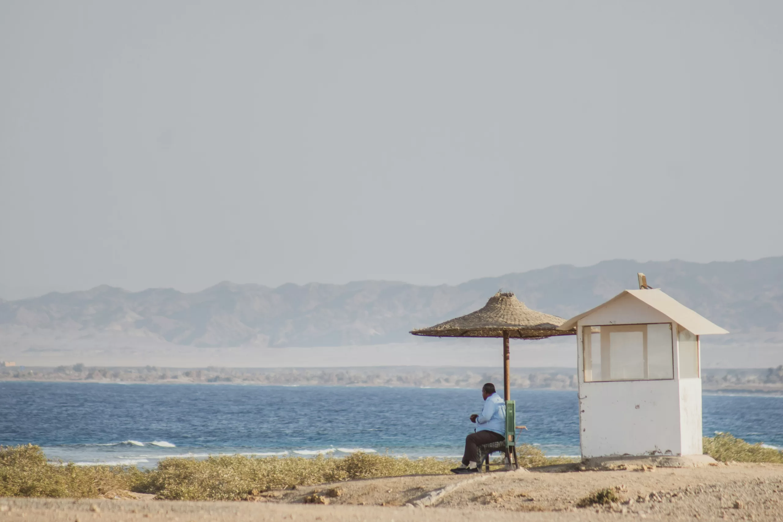 Man sitting beneath a small shade structure beside the sea, with distant mountains visible across the water under a hazy sky