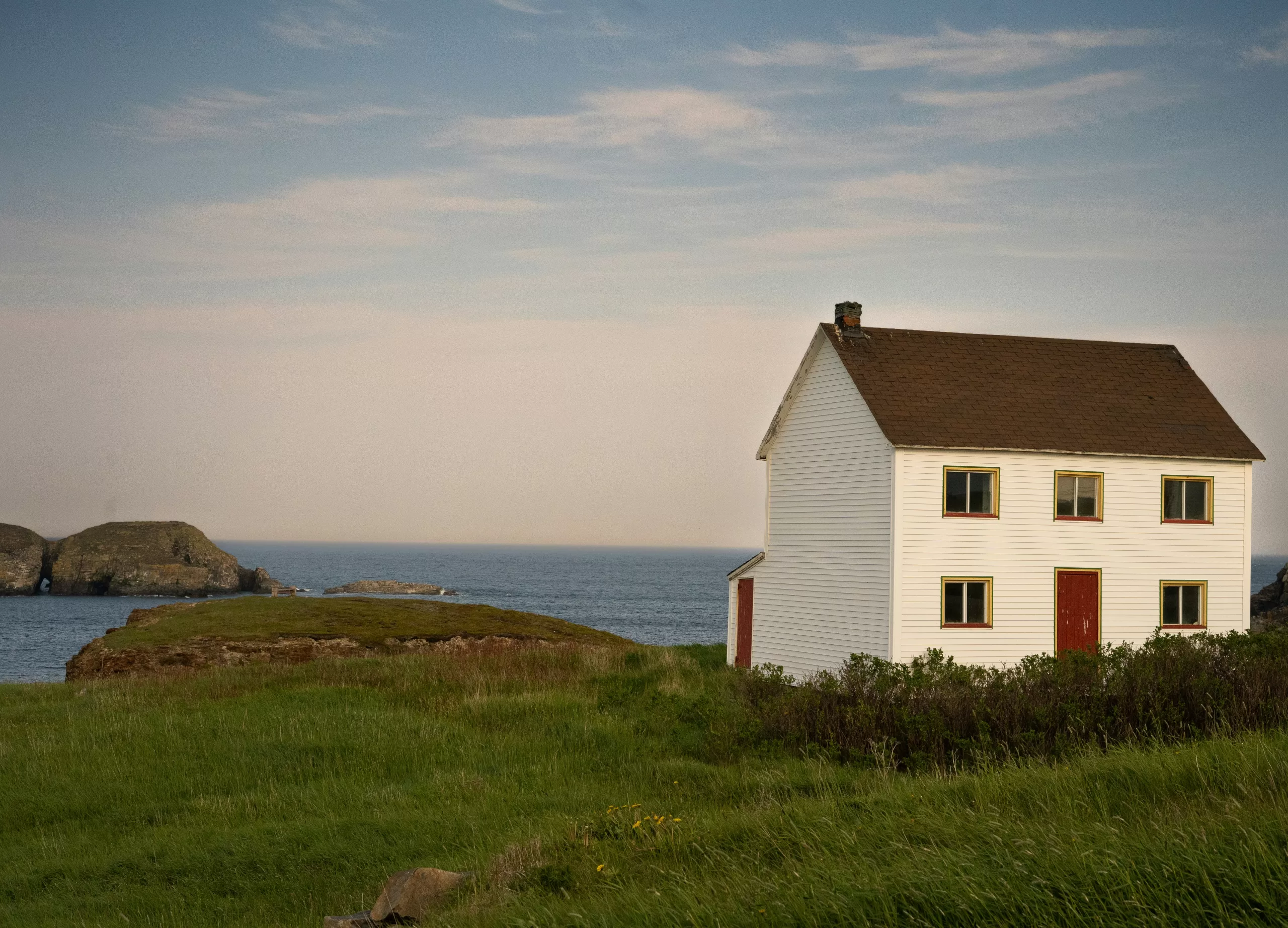 White house standing on a grassy coastal bluff overlooking the ocean, with rocky outcrops and open sky in the background