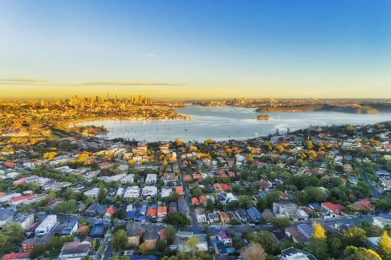 Aerial view of Sydney Harbor and surrounding suburbs at sunset, illustrating Australia’s housing affordability debate