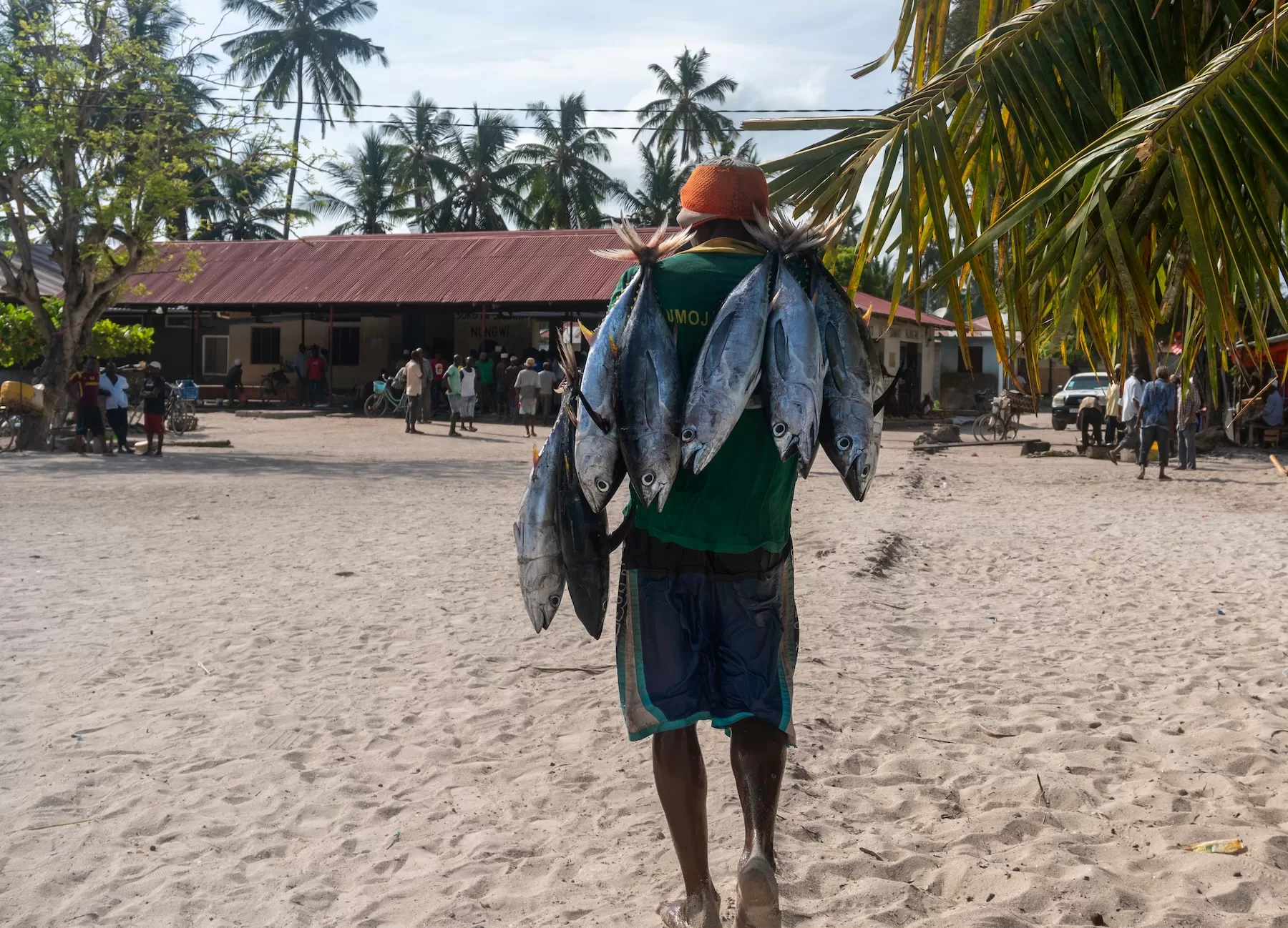 A man walks across a sandy village square carrying several large fish over his shoulders in a coastal Zanzibar community.