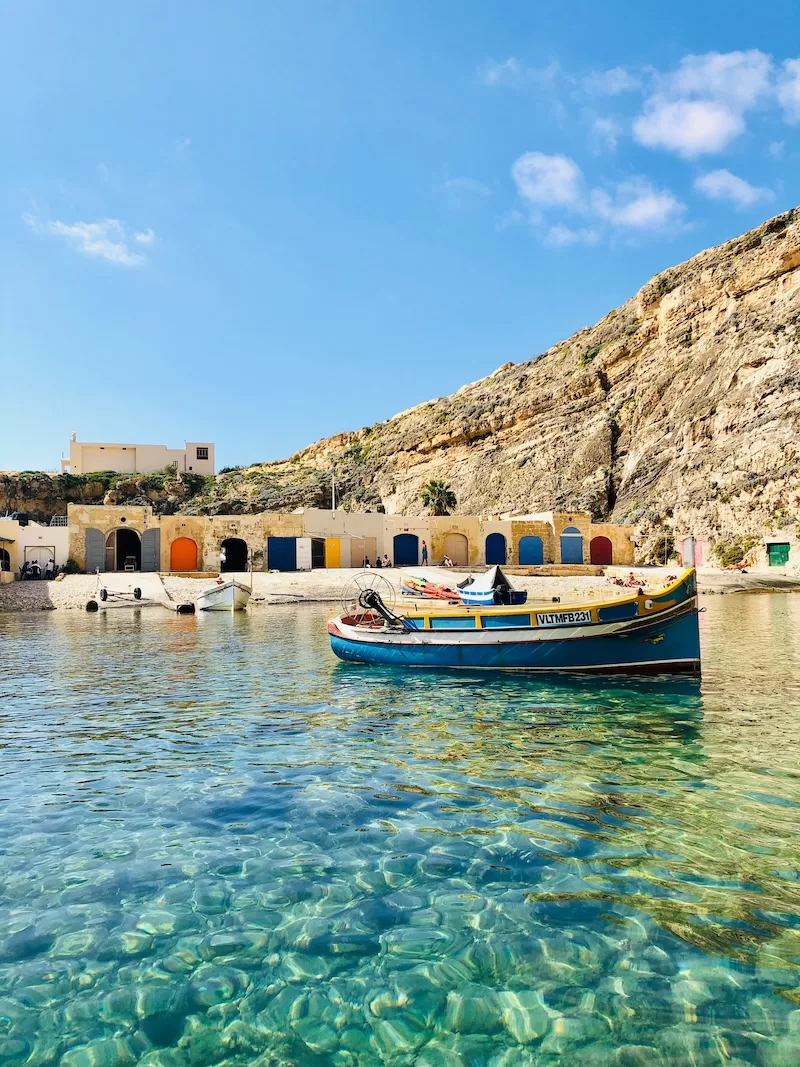Small Mediterranean harbor with fishing boat and colorful boathouses beneath rocky cliffs in Malta