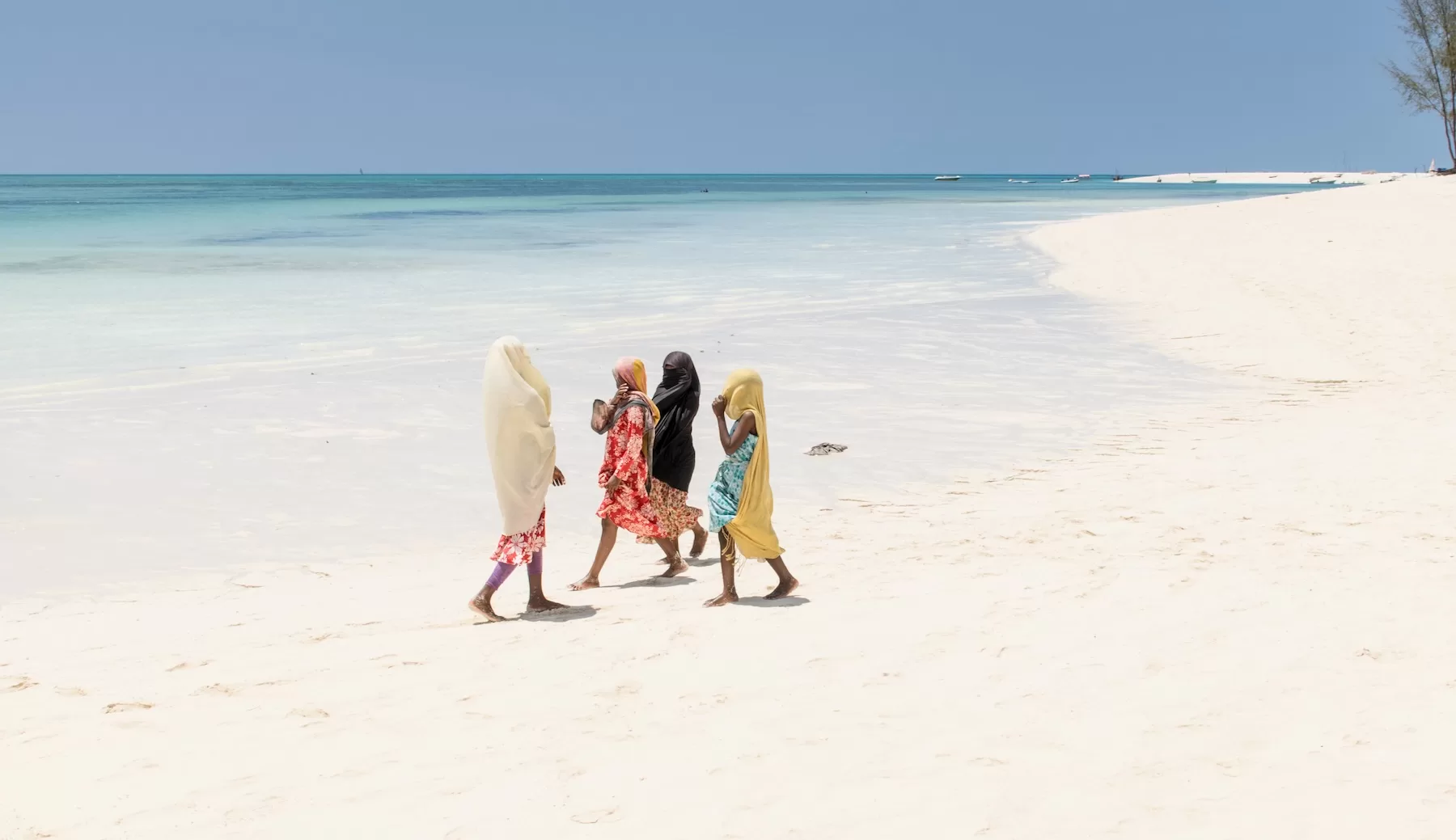 Three women walk along a broad white beach in Zanzibar beside shallow clear blue water.