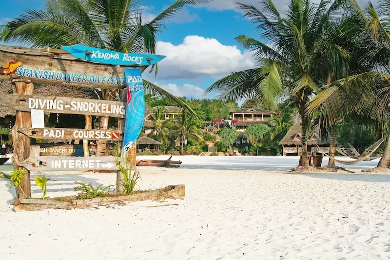 Rustic beach sign at Kendwa Rocks in Zanzibar points to diving, snorkeling, PADI courses, and internet near palm trees and beach lodges.