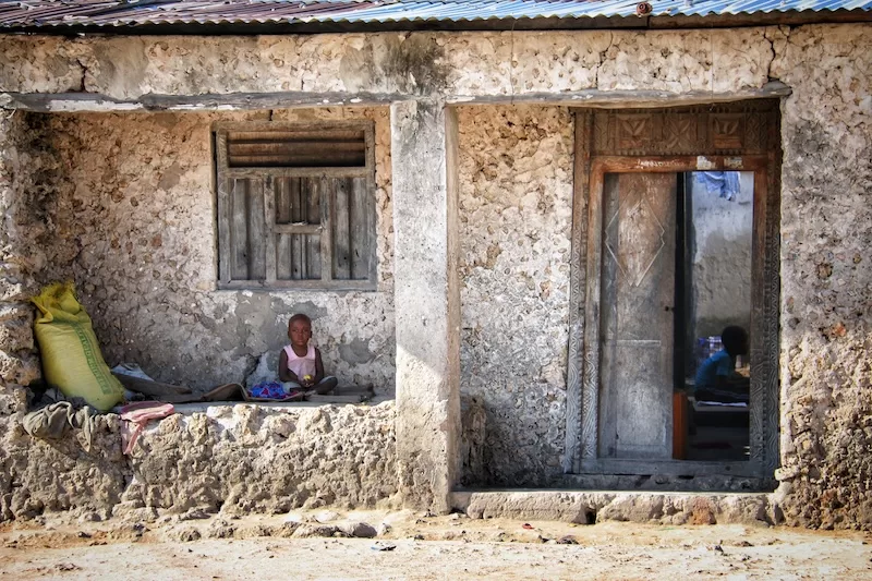 A young child sits outside a weathered coral-stone house with carved wooden door and window in Zanzibar.