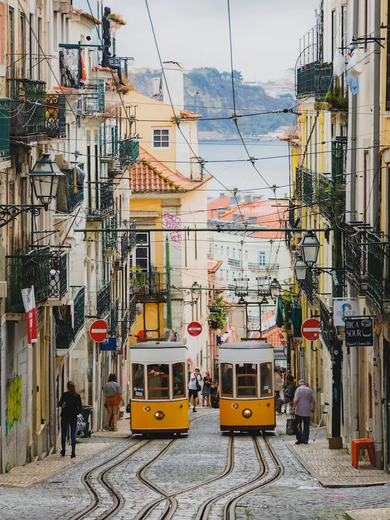 Yellow trams passing through a narrow Lisbon street with tiled buildings and river views.