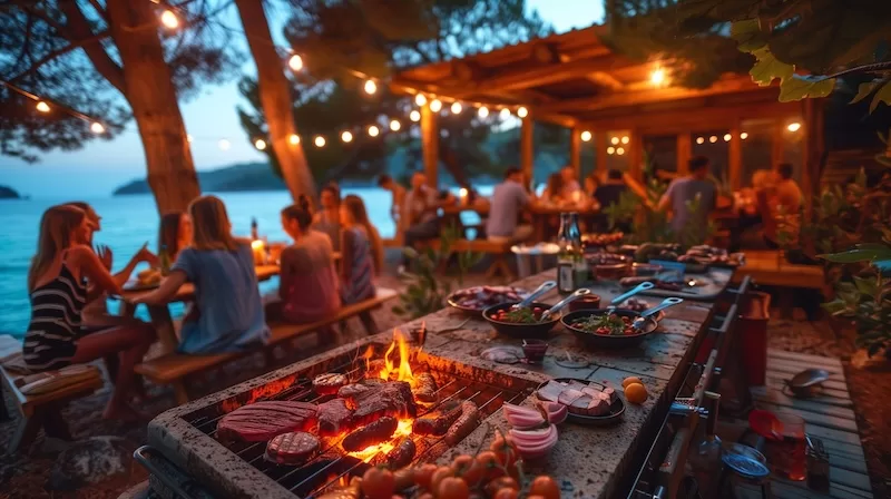 People gather around a beachside barbecue and dinner setup at sunset in Zanzibar with food grilling beside the water.