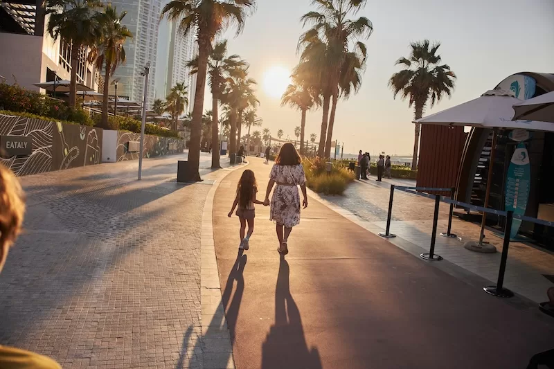 Woman and child walking along a palm-lined waterfront promenade in a modern Gulf city