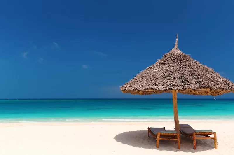 A thatched beach umbrella and wooden loungers sit on a quiet white-sand Zanzibar beach beside vivid turquoise water.