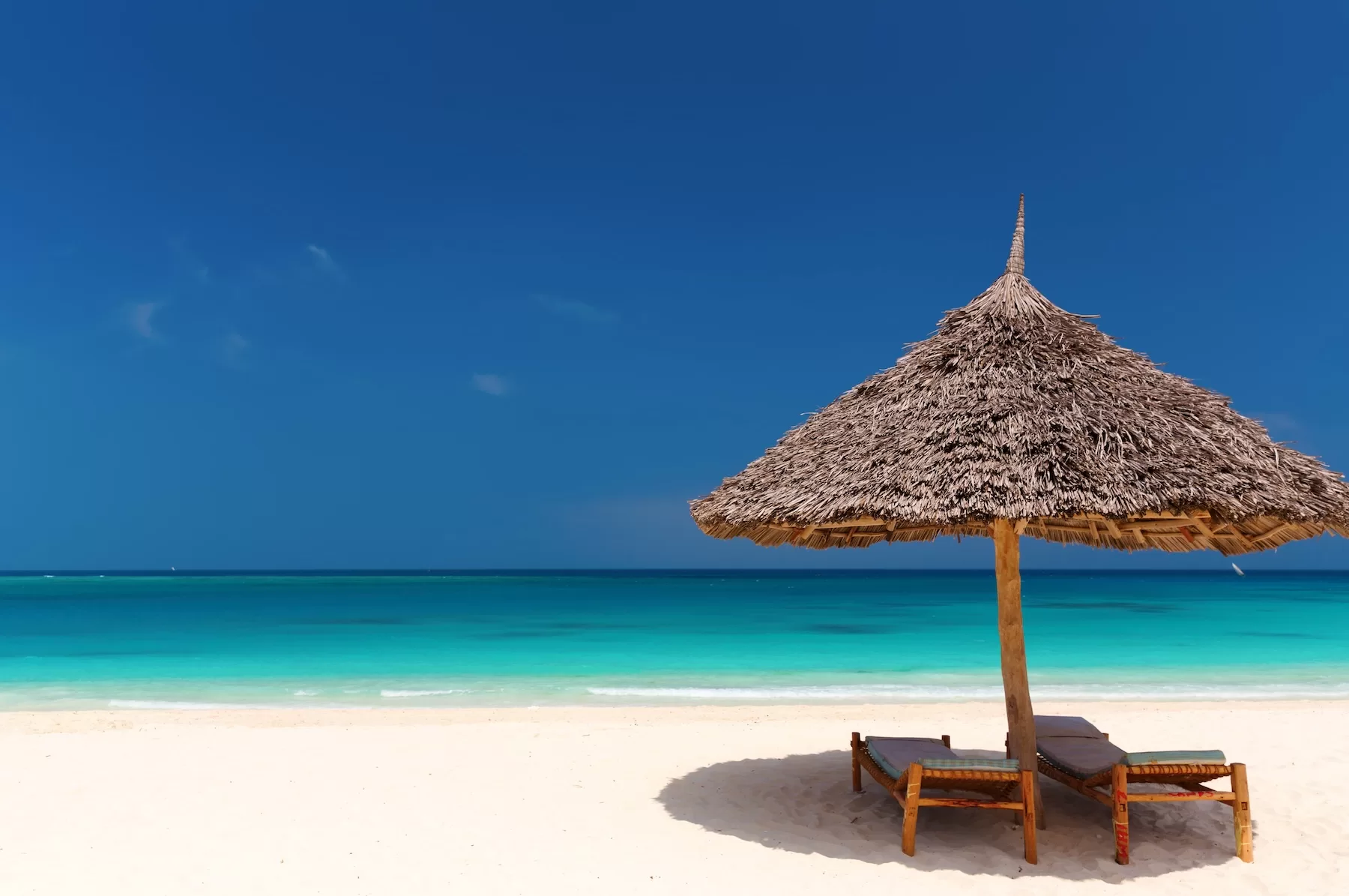 A thatched beach umbrella and wooden loungers sit on a quiet white-sand Zanzibar beach beside vivid turquoise water.