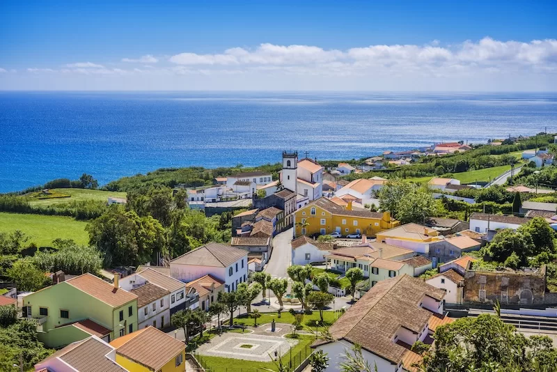 Coastal village in the Azores with colorful houses, green fields, and the Atlantic Ocean in the background