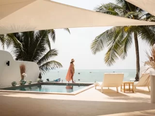 Woman in a sun hat walking beside an infinity pool overlooking the ocean under palm trees