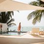 Woman in a sun hat walking beside an infinity pool overlooking the ocean under palm trees
