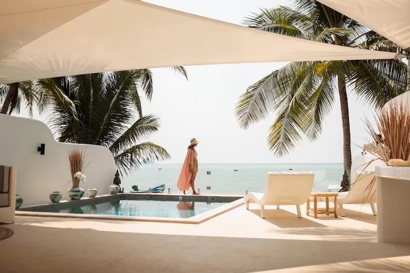 Woman in a sun hat walking beside an infinity pool overlooking the ocean under palm trees