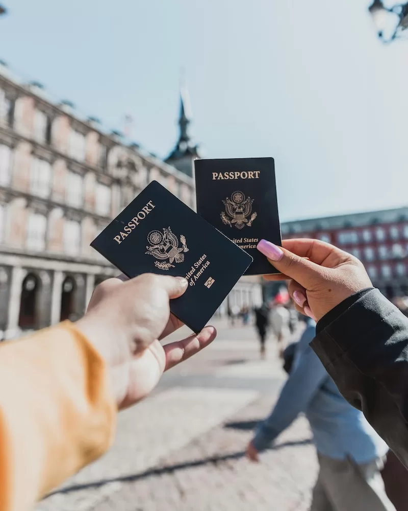 Two hands holding US passports in a sunlit plaza in Madrid