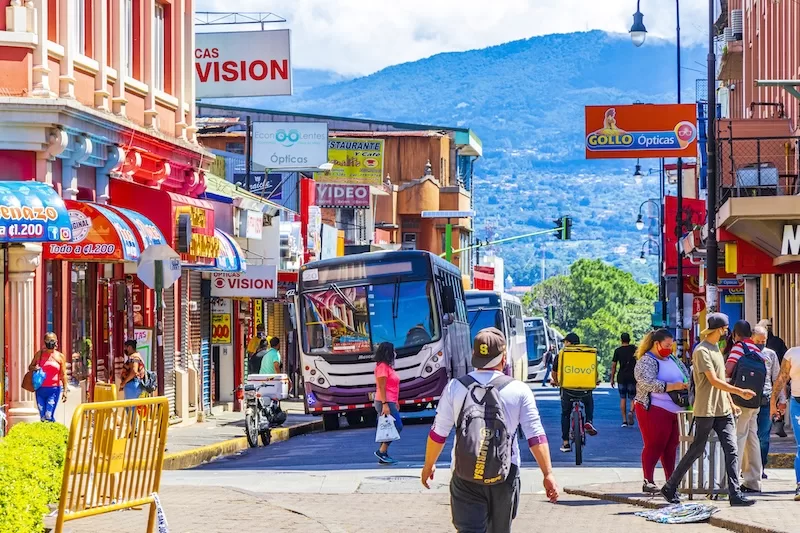 Busy Costa Rican city street with buses, pedestrians, storefronts, and mountains in the background