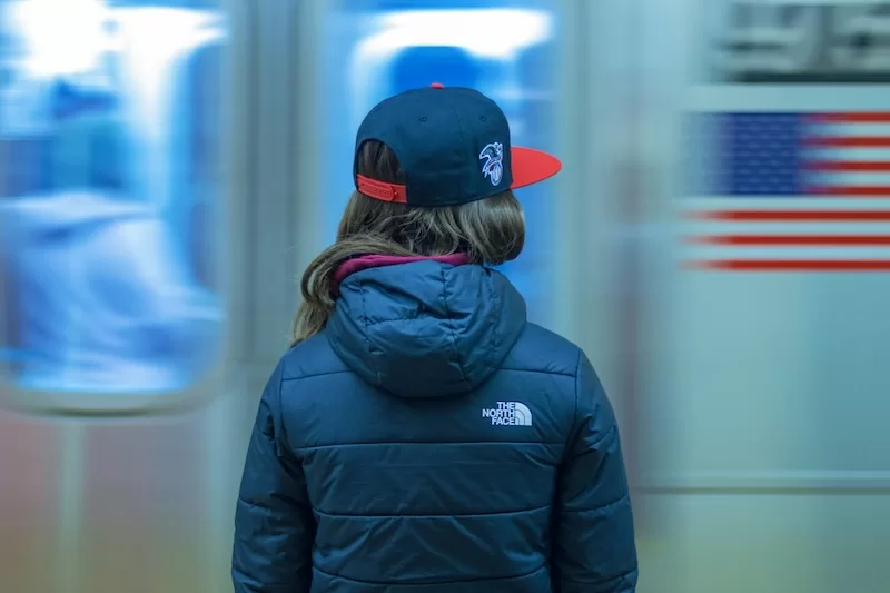Person viewed from behind wearing a cap and puffer jacket with an American flag blurred in the background