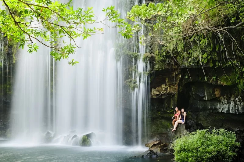 Two travelers sitting beside a waterfall surrounded by dense green forest in Costa Rica