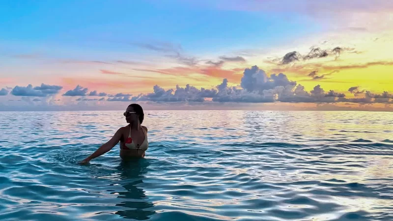 Woman standing in the ocean at sunset with pastel-colored clouds on the horizon