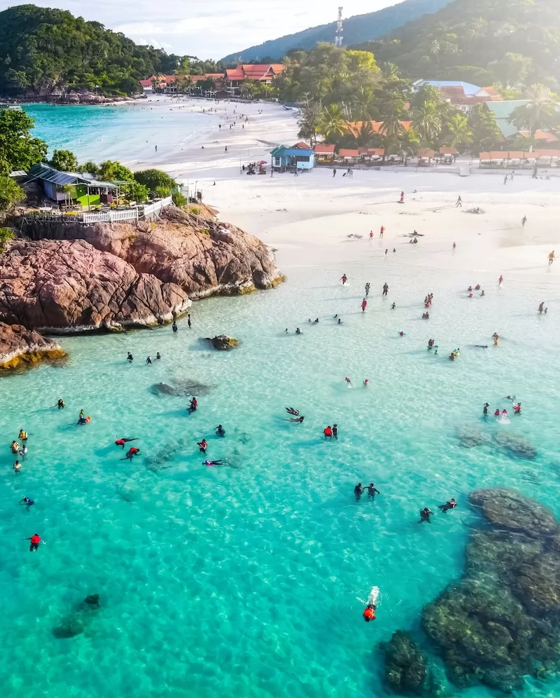 Aerial view of swimmers in shallow turquoise water off a white sand beach in Malaysia
