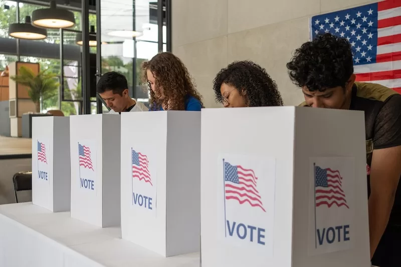 People filling out ballots at voting booths in front of an American flag