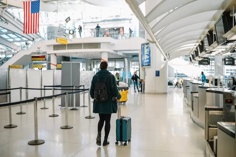 Traveler with a suitcase walking through an airport terminal beneath a hanging American flag