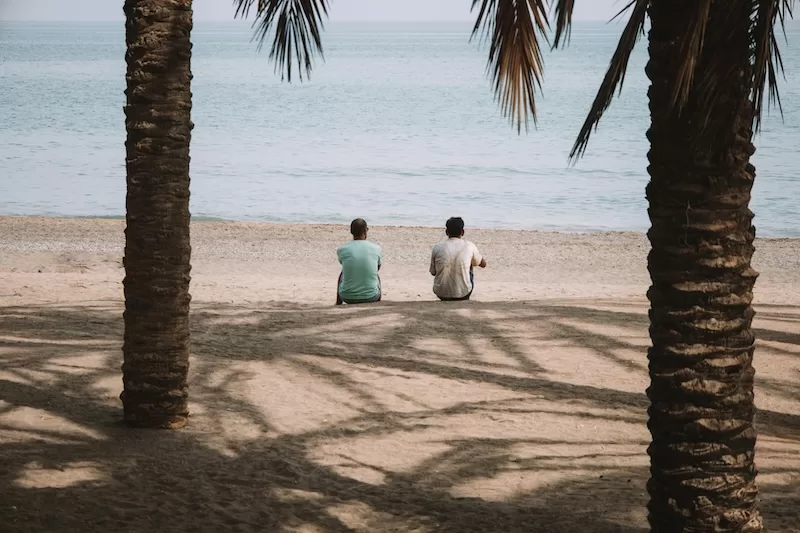 Two people sitting beneath palm trees on a quiet beach in southern Spain