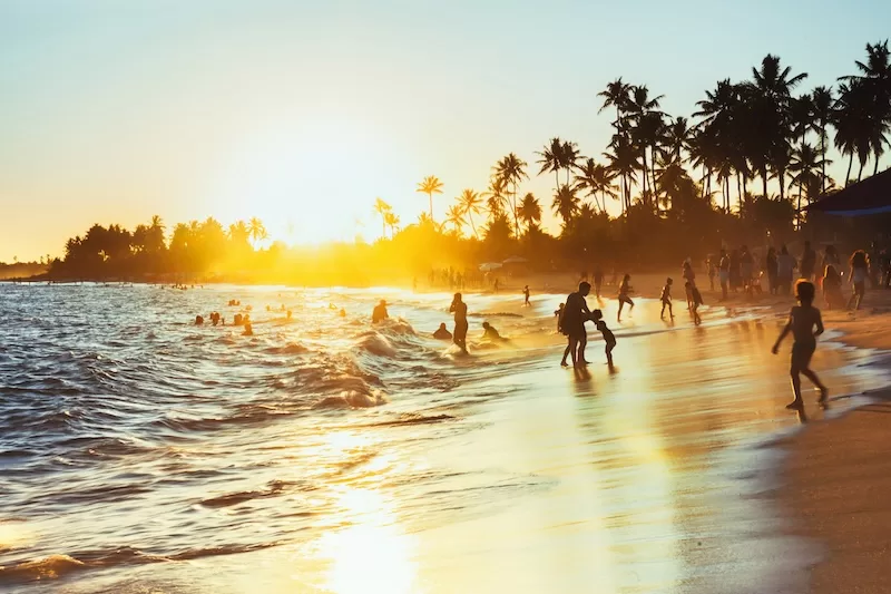 A crowded Brazilian beach at sunset, families playing in the surf under palm trees: where the story of moving from Brazil to Europe begins
