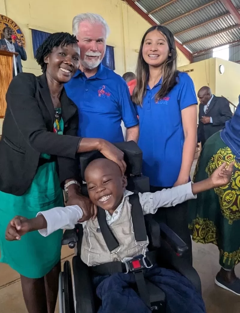 A young boy beams from a specialty wheelchair beside his mother and volunteers at a distribution.