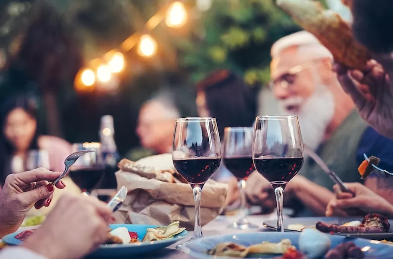 Glasses of red wine at a long outdoor dinner under string lights.