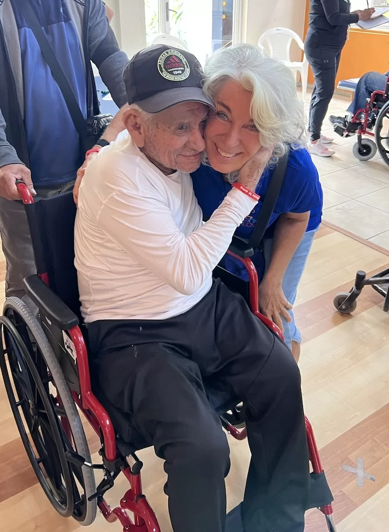 An elderly man in a new wheelchair embraces a volunteer.