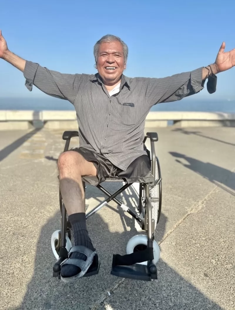 A man raises both arms in celebration, sitting in his new wheelchair on a seaside promenade.