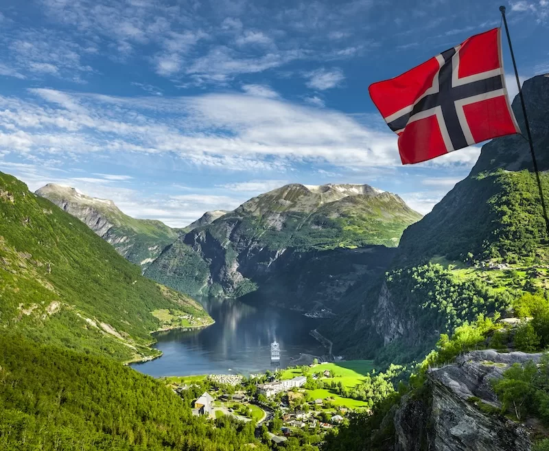 A Norwegian flag above Geirangerfjord, with a cruise ship moored at the village far below