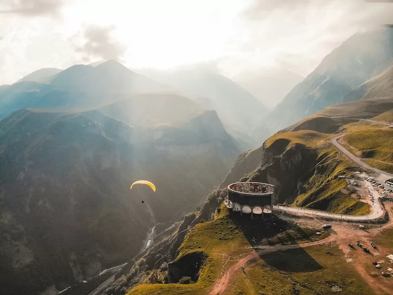 A paraglider drifts past a circular mountain viewpoint high above a gorge.