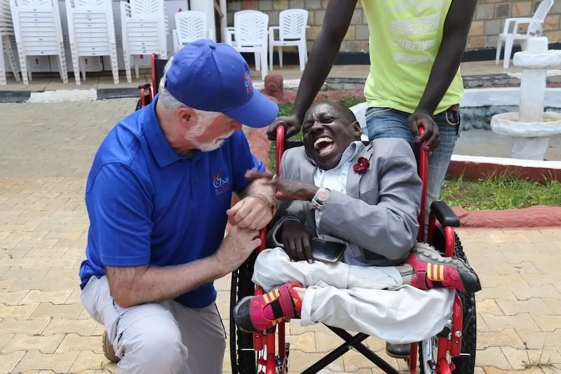 A volunteer kneels beside a young man laughing in his new wheelchair.