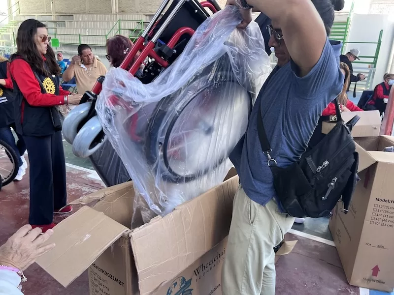 A volunteer lifts a new wheelchair from its shipping box while Rotary members assist.