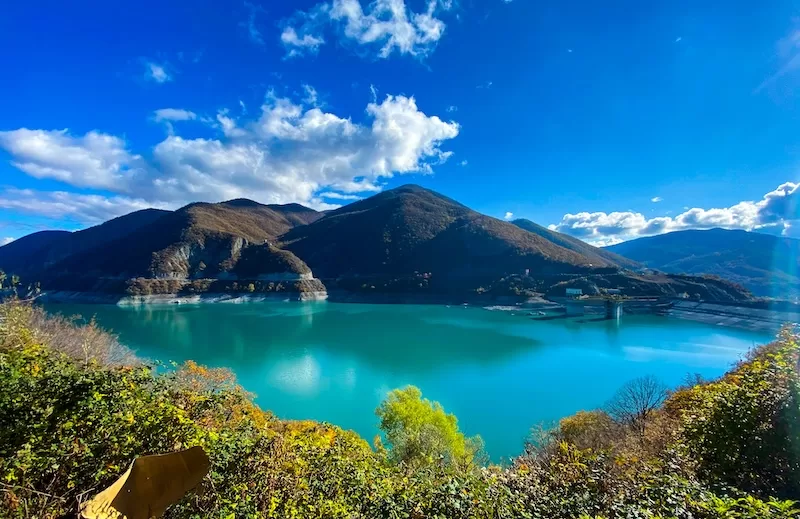 A turquoise mountain reservoir under a bright blue sky, ringed by autumn hillsides.