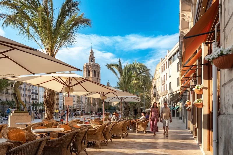 A café terrace on a sunlit Valencia square, the Miguelete tower behind.
