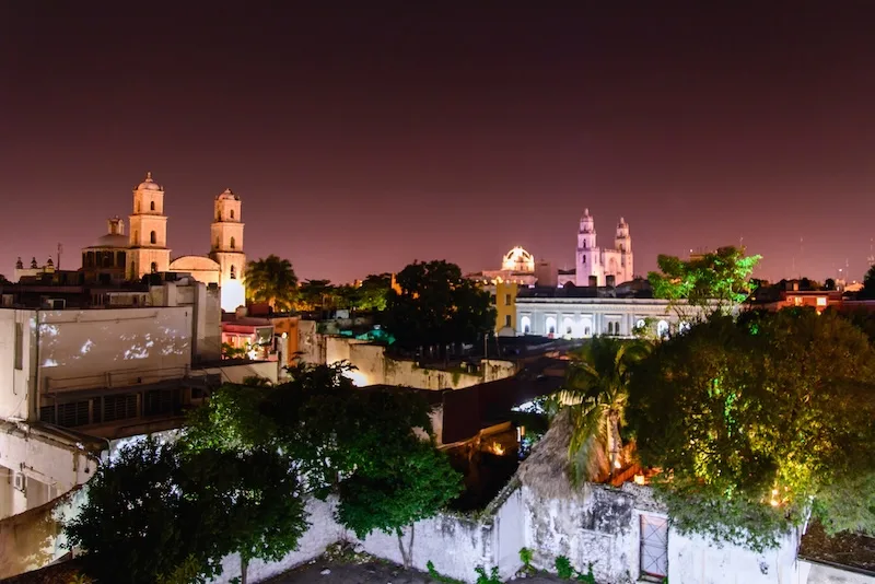 Floodlit colonial church towers rise over Mérida’s rooftops at night.